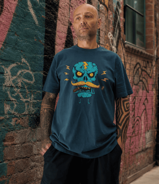 A man wearing a dark teal graphic t-shirt with a cartoon skull design, standing against a graffiti-covered brick wall.