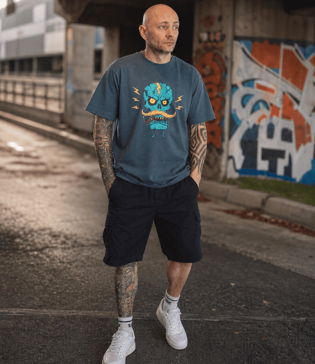 A man wearing a slate blue graphic t-shirt with a cartoon monster design, black cargo shorts, and white sneakers, standing on an urban street with graffiti walls in the background.